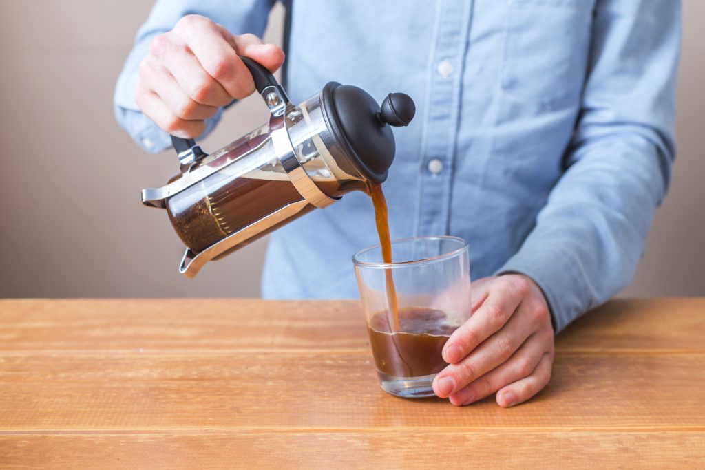 seorang lelaki berkemeja biru menuangkan kopi hitam hasil seduhannya dari french press ke dalam gelas